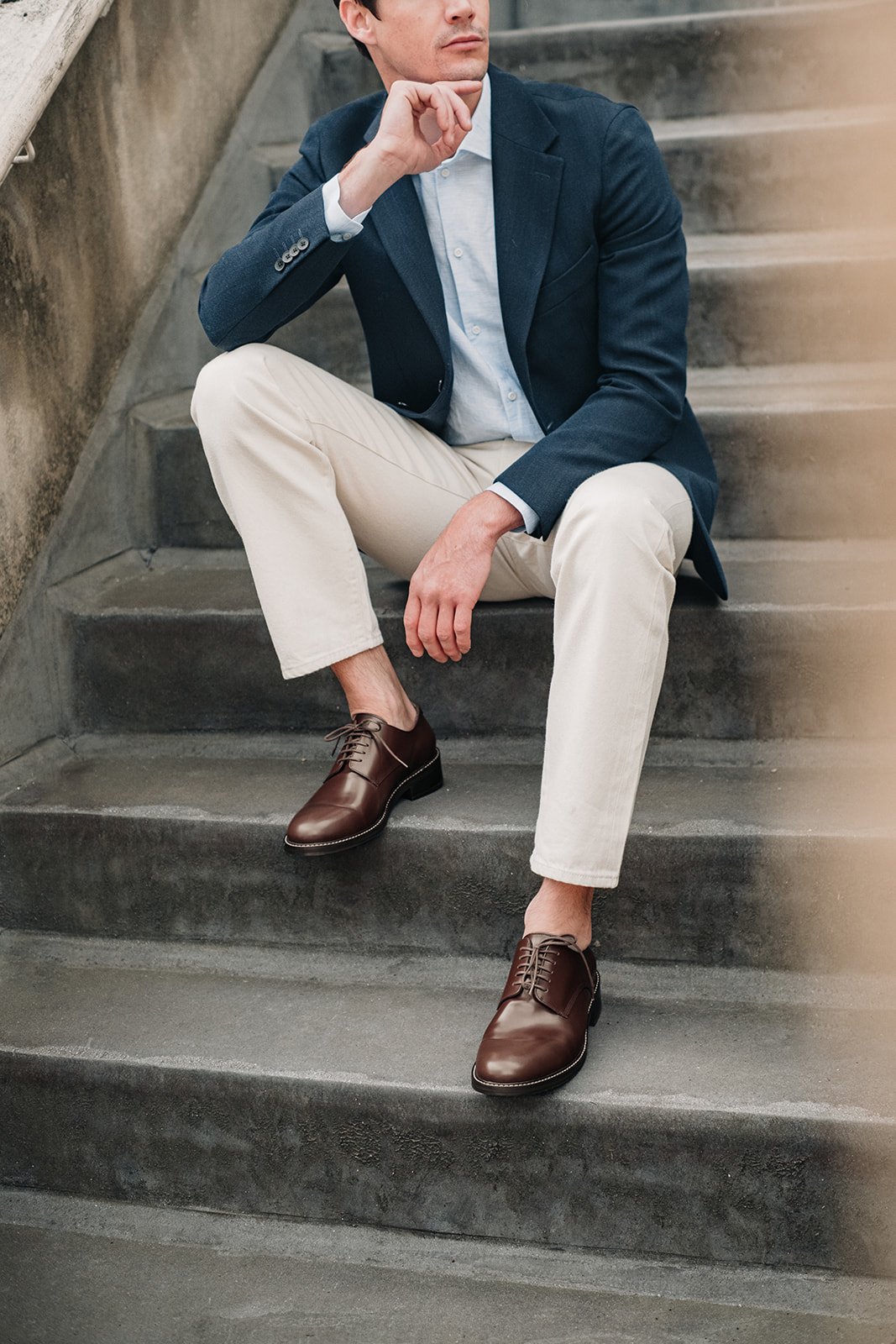 Modèle masculin assis sur des escaliers à Londres, portant des chaussures en cuir marron et un blazer bleu marine.