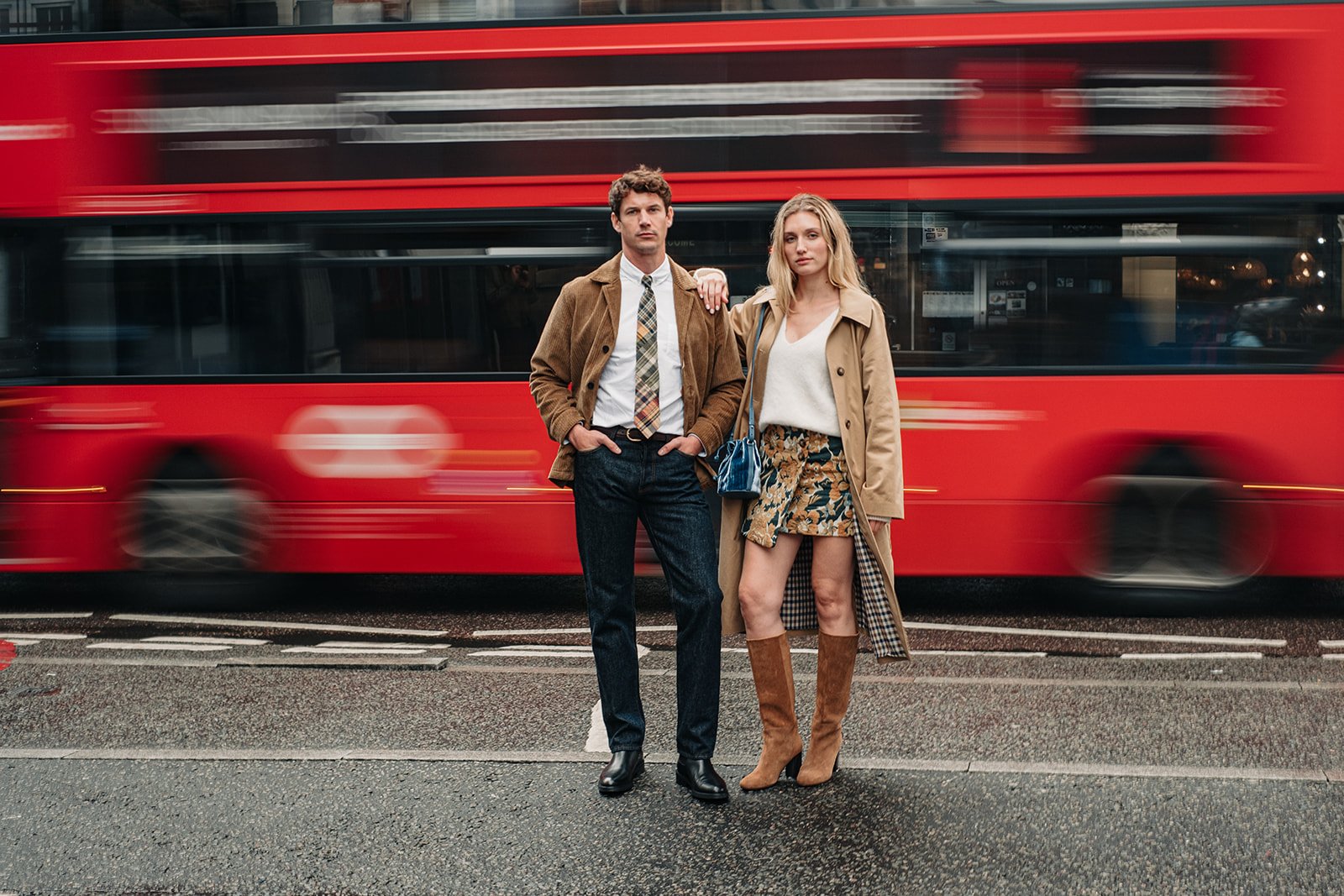 Couple posé devant un bus rouge londonien en mouvement, portant des vêtements et chaussures Pied de Biche.